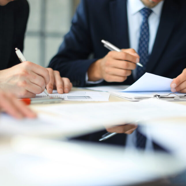 Sitting close in a table, three people in business attire write with pen on paper documents — first opinion coverage from STAT