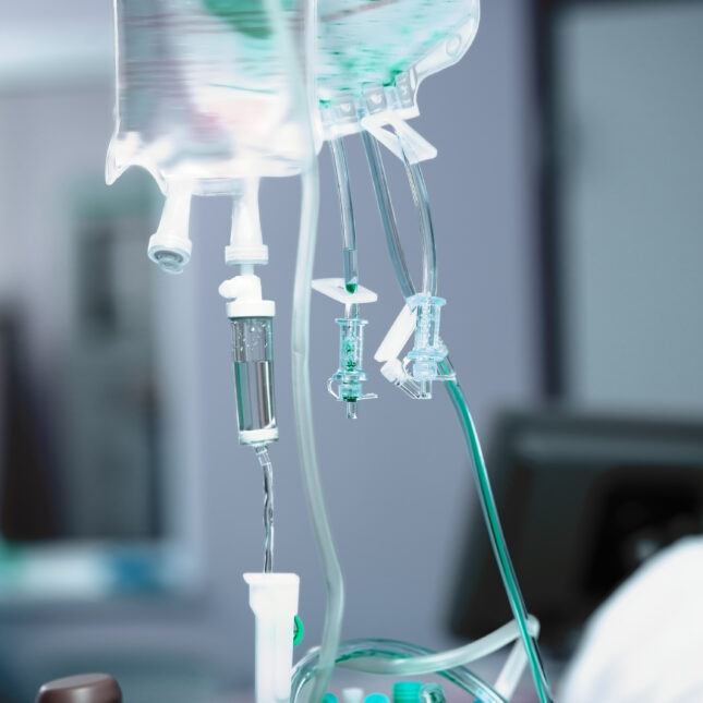 Two Intravenous drip bags hung next to each other, with the right one shining in slightly teal light — health coverage from STAT