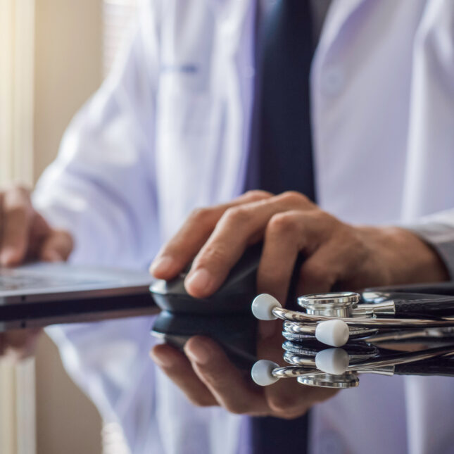 A doctor holds a wireless mouse in front of a laptop and next to a stethoscope — First Opinion coverage from STAT