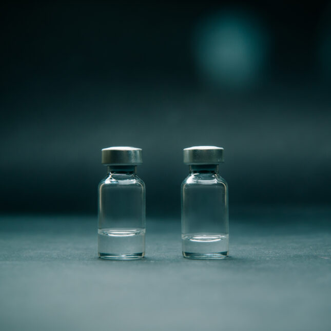 Two vials stand next to each other in a dark teal environment. The left one contains 1/3 vial of liquid and the left one contains slightly less — first opinion coverage from STAT