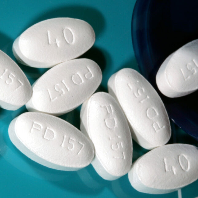 Nine tablets of Lipitor in white oval shape comes out a dark blue pill bottle from the right — health coverage from STAT