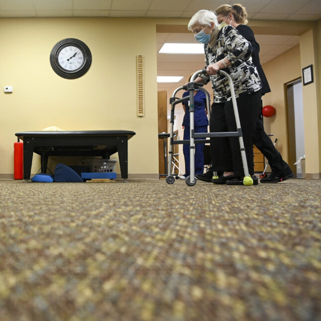 On the right side of a frame, a nurse supports a senior as she moves with a folding walker — first opinion coverage from STAT