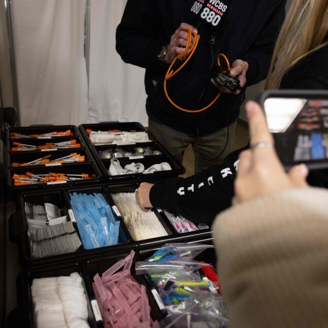 A person picks up a piece of medical equipment to show media at a supervised drug injection site. Another person records the organized boxes that contain a variety of tools, such as syringes, alcohol wipes, and bandages, with their phone. — First Opinion coverage from STAT