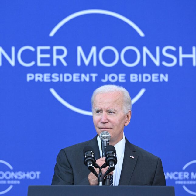 US President Joe Biden holds a microphone while speaking at a podium in front of a blue backdrop. The backdrop displays the title "CANCER MOONSHOT" above a line of smaller text that reads "PRESIDENT JOE BIDEN," both lines partially enclosed in a circular border — health coverage from STAT