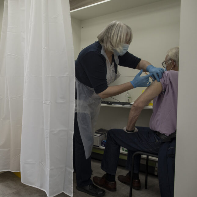 A person gets vaccinated on their left arm behind a curtain as two people wait in line, sitting outside the room — health coverage from STAT