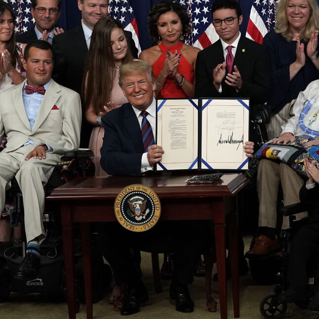 Former U.S. President Donald Trump shows his signing of the "Right to Try" Act, surrounded by a group of people, some sit in power wheelchairs, some standing up, and some clapping — first opinion coverage from STAT