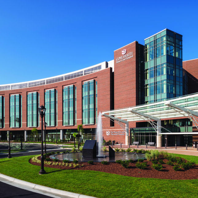 A hospital building with a brick exterior, labeled "CONE HEALTH Moses Cone Hospital" on the top, stands next to its driveway— hospitals coverage from STAT