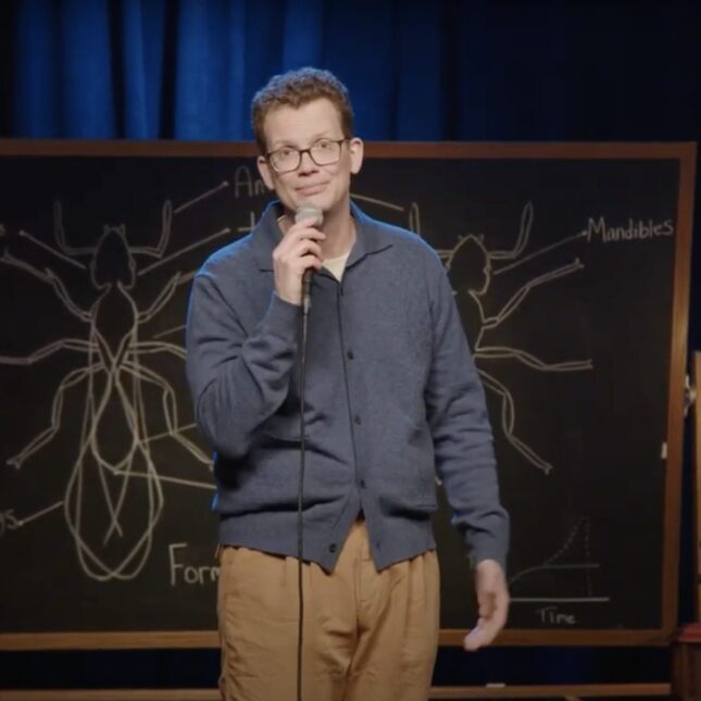 Hank Green on stage holding a microphone with a chalkboard behind him. -- coverage from STAT