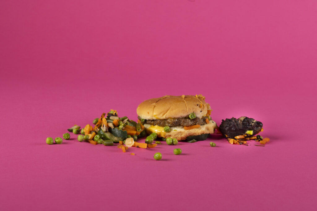 A studio photograph of Homestyle Direct's Homestyle Cheeseburger meal costs $9.95 and comes with "Scandinavian vegetables" and "brownie bites." on a bright fuschia backdrop