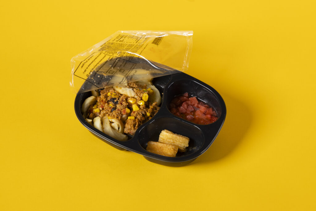 A studio photograph of Homestyle Direct's "Idaho Nachos" frozen meal with churro bites and strawberries. Photographed on a bright yellow backdrop.